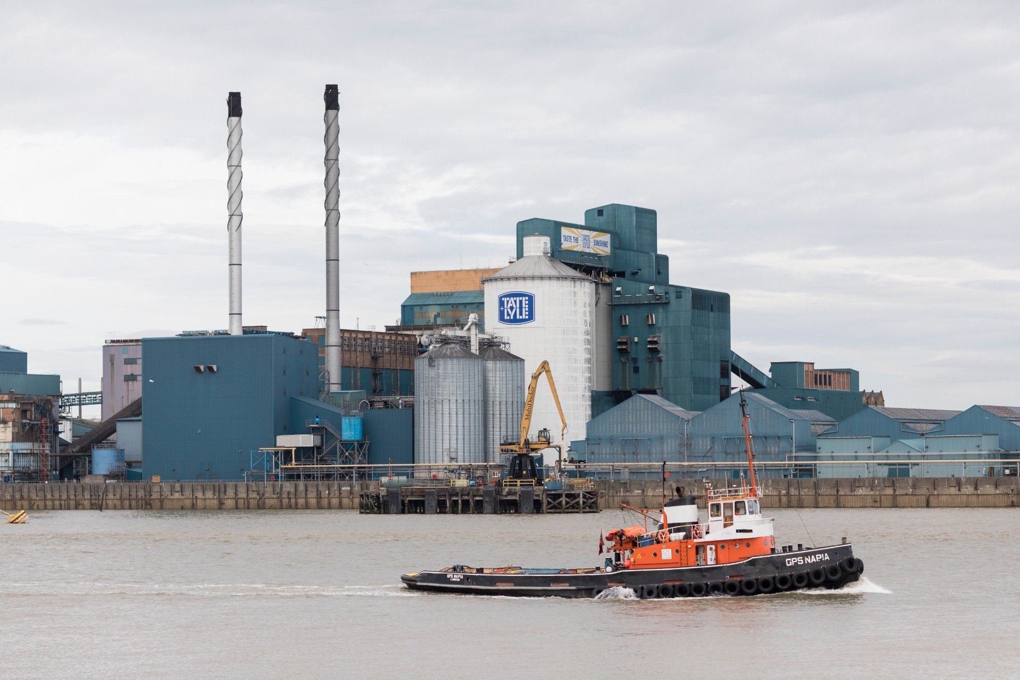 Tate & Lyle building exterior from the river with a boat in the foreground and big chimneys visible