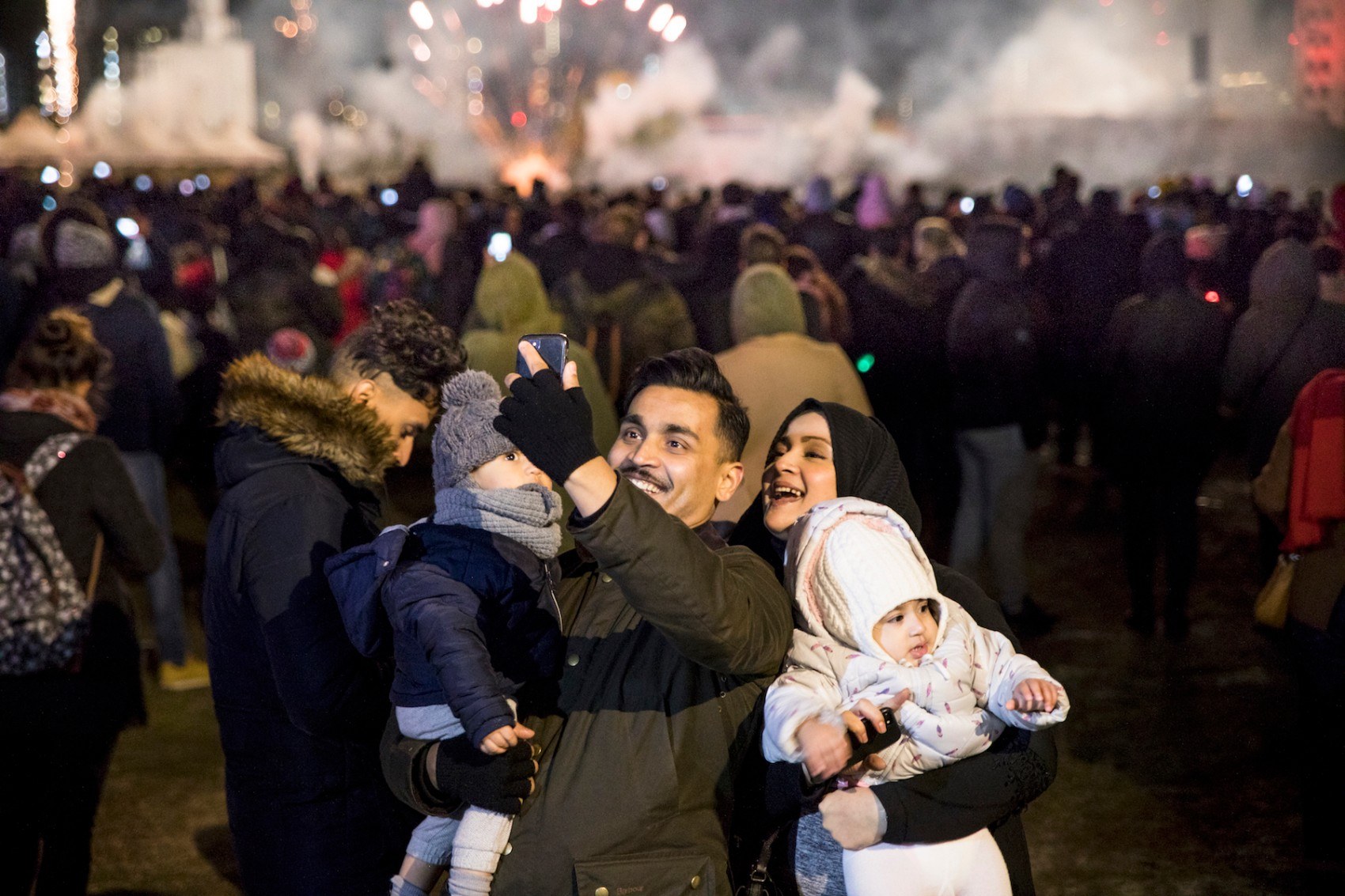 Man and woman holding a child each taking a selfie with fireworks in the background