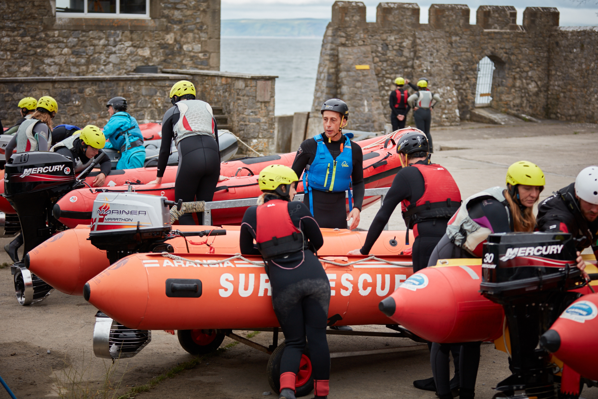 A group of people in wetsuits surrounding a group of four lifeboats
