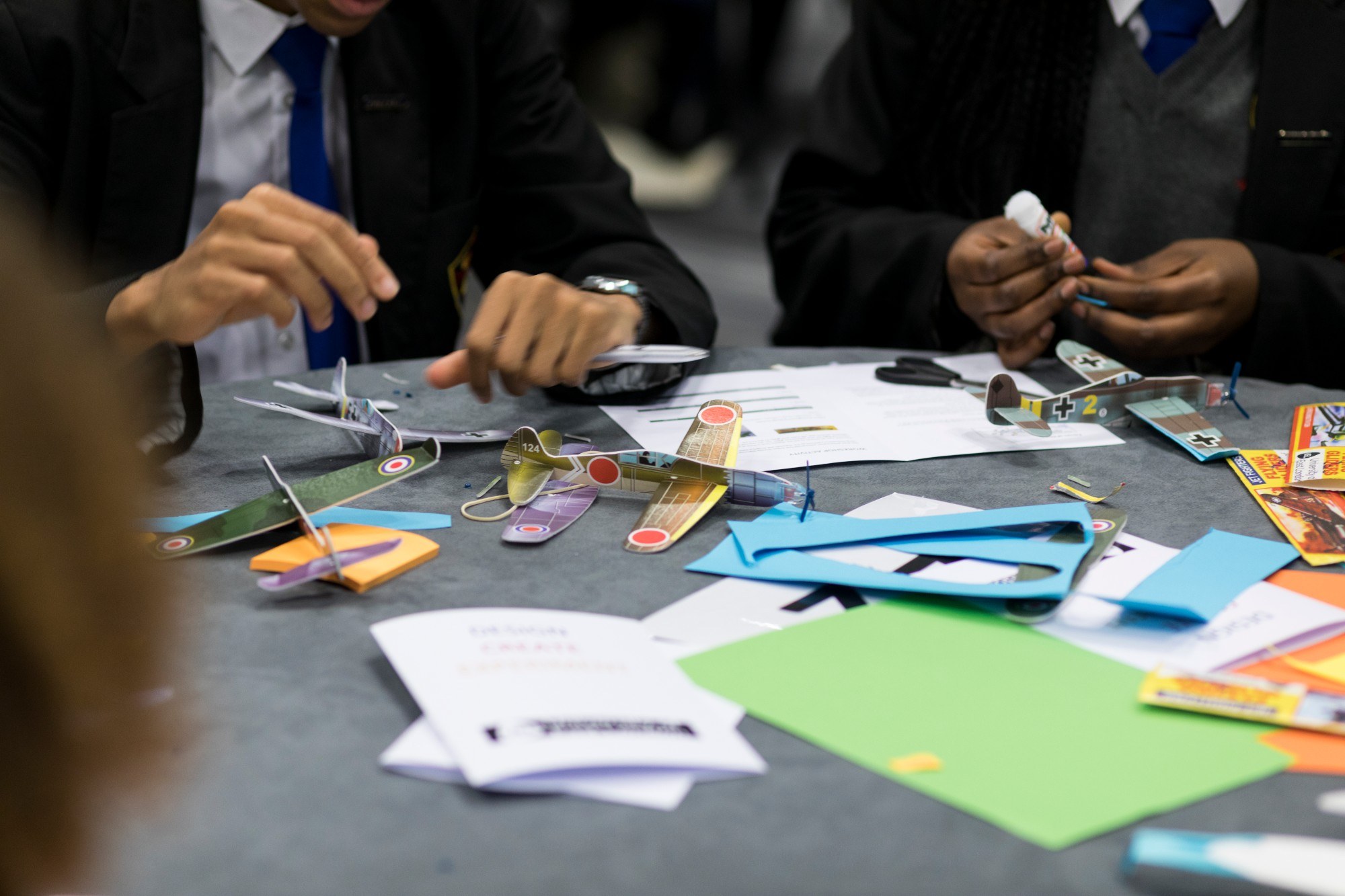 Small model planes and coloured paper on a table, with children working on them